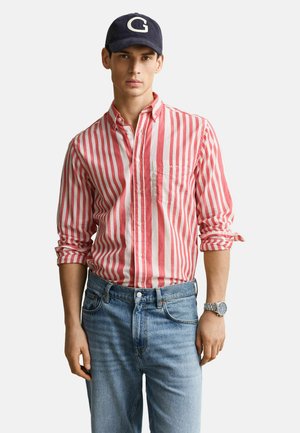 Mann mit marineblauer Kappe, rot-weiß gestreiftem Hemd mit aufgekrempelten Ärmeln, hellblauer Jeans und einer silbernen Armbanduhr.