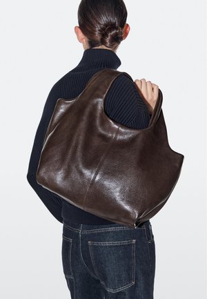 Personne avec un chignon foncé portant un pull noir et un jean, tenant un grand sac cabas en cuir marron foncé sur l'épaule.