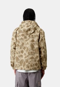 Giacca camouflage in tonalità beige, con cappuccio, polsini elastici e vestibilità leggermente ampia. Realizzata in un materiale liscio e resistente all'acqua.