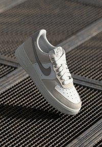 Nike Air Force 1 toss hall halli ja valge. Omab nahast ja seemisnahast materjali, aukudega varvast ja tekstuurse kummipõhjaga, millel on paelad.