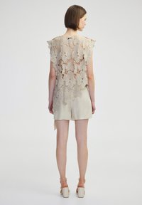 Un haut en dentelle présentant des motifs floraux complexes en beige, avec une coupe ample et des manches fringées, associé à un short léger et des talons épais.