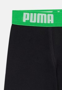Crne tajice s zelenim pojasom na kojem je bijeli tekst "PUMA". Tkanina izgleda glatko s finim šavovima uz rubove.