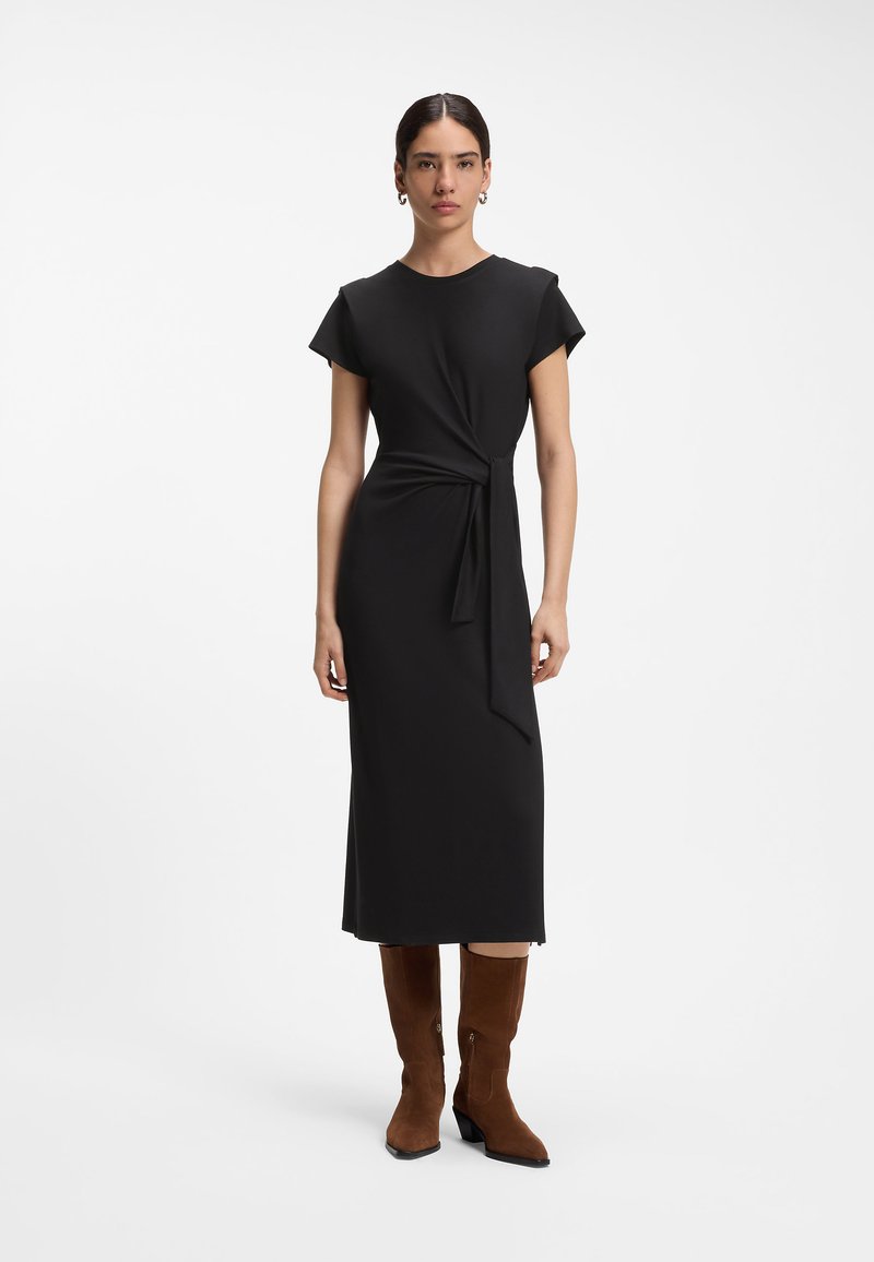 Femme aux cheveux foncés attachés, portant une robe midi noire à manches courtes avec un nœud sur le côté et des bottes en daim marron montant jusqu'au milieu du mollet, debout sur fond blanc.