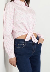 Vit skjorta med rosa geometriskt mönster, knuten i midjan, parat med mörkblå jeans. Långa ärmar med knappmanchetter.