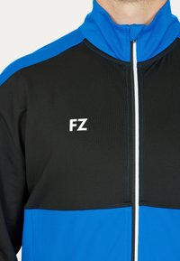 Blå och svart zip-jacka med hög krage, som har vit sömmar och "FZ"-logotyp i vitt. Texturerad tyg med färgblockeringsdesign.