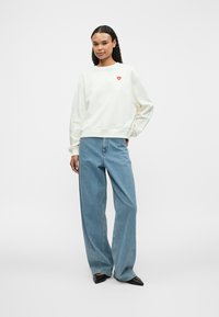 Light cream sweatshirt with a red heart logo, relaxed fit, ribbed cuffs, paired with light blue high-waisted wide-leg jeans, and black pointed flats.