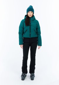 Moteris vilkinti mėlyną pūkinę striukę, atitinkančią megztą kepurę, juodus sniego kelnes ir juodus slidinėjimo batus, stovinti ant balto fono.