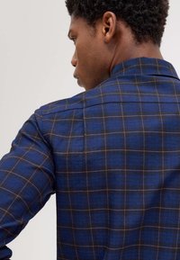 Camicia a quadri blu con tessuto testurizzato, linee marroni e nere, colletto e design con bottoni visibile dal retro.