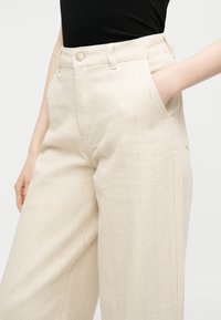 Άτομο που φοράει υφασμάτινο παντελόνι κρεμ χρώματος με ψηλή μέση, κάθετες ραφές και τσέπες μπροστά, σε συνδυασμό με μαύρο μπλουζάκι.