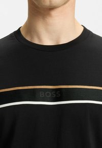 Black T-shirt with a round neck, featuring horizontal stripes in gold, green, and white. Logo "BOSS" displayed on the front. Textured fabric.