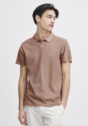 Polo shirt marron à manches courtes avec un col, en tissu texturé, affichant un logo subtil sur la poitrine. Porté avec un pantalon de couleur claire.