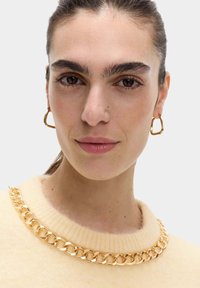 Gebreide trui in zacht geel met een textuur van een gouden kettinghalslijn, gecombineerd met gouden creolen, met een gladde, gepolijste afwerking.