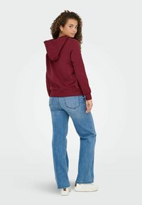 Bordeaux hættebluse med snøretiltræk, blød tekstur og ribbede manchetter, parret med lyseblå denimjeans og hvide sneakers.