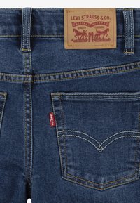 Vue arrière d'un jean Levi's bleu montrant la patch en cuir de la marque et l'étiquette en tissu rouge Levi's sur la poche arrière droite avec des détails de couture jaune.