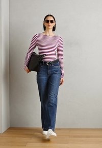 Gestreiftes langärmliges Oberteil in Lila und Weiß, hochgeschnittene blaue Jeans, schwarze Tasche und weiße Sneaker. Model läuft auf Holzfußboden vor einer grauen Wand.