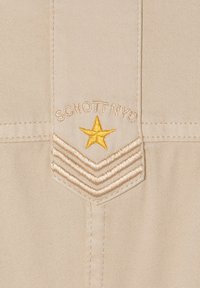 Beige Stoffpatch mit "SCHOTT NYC" in Braun bestickt und einem gelben Stern über drei chevronartigen Linien. Glatte Textur, detaillierte Stickerei.