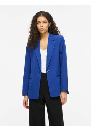 Göndör hajú nő kék blézert visel fehér felső és fekete nadrág fölött, egyszerű fehér háttér előtt állva.