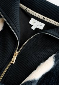 Mustaline tõmblukuga jakk, millel on soonikmaterjal, kuldne tõmblukk ja pehme kunstkarvast kaelus. Siseetiketil on kirjas "b by Ted Baker London."