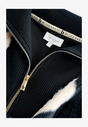 Juodas užtrauktukas striukė, pasiūta iš banguotos medžiagos, su auksiniu užtrauktuku ir minkštu dirbtinio kailio apykakle. Vidinė etiketė skaito "b by Ted Baker London."