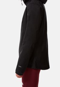 Person, der eine schwarze wasserdichte Jacke mit Kapuze trägt, von der Seite gezeigt, kombiniert mit burgunderfarbenen Leggings.