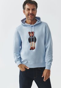 Sudadera azul claro con un gráfico de oso, combinada con un suéter negro y pantalones cortos beige. Tejido texturizado con cordones y puños acanalados.