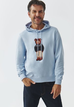 Sudadera azul claro con un gráfico de oso, combinada con un suéter negro y pantalones cortos beige. Tejido texturizado con cordones y puños acanalados.