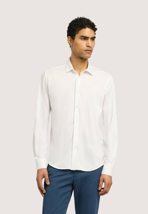 Mann trägt ein langärmeliges weißes Hemd mit Knöpfen und blaue Hose, schaut leicht nach rechts vor einem schlichten Hintergrund.