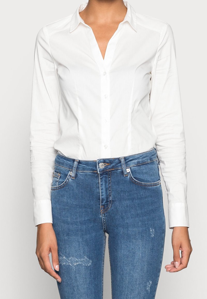 Woman wearing a fitted white long-sleeve button-up shirt and blue denim jeans with slight distressing on thighs and pockets.