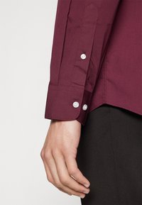 Camisa burdeos con un puño estructurado y tres botones blancos. Textura de tela suave; diseño ajustado con costuras adaptadas.