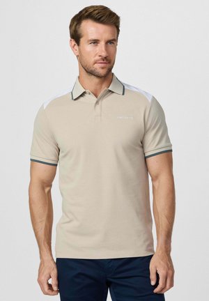 Hombre vestido con una camiseta polo beige con paneles blancos en los hombros y pantalones cortos azul marino, de pie frente a un fondo liso y claro.