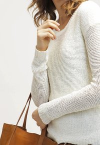 Biała dziergana sweter z długimi rękawami i teksturowanym wzorem. W drugiej ręce trzymana jest brązowa skórzana torba.