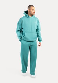 Sweatshirt capuche turquoise avec une poche avant, associé à un pantalon de survêtement assorti. Le matériau semble doux et texturé. Des baskets blanches complètent la tenue.