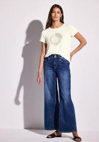 Lys gul t-shirt med cirkulært grafik, parret med løse blå jeans og sorte slidesandaler. Glatte stofteksturer og afslappet design.