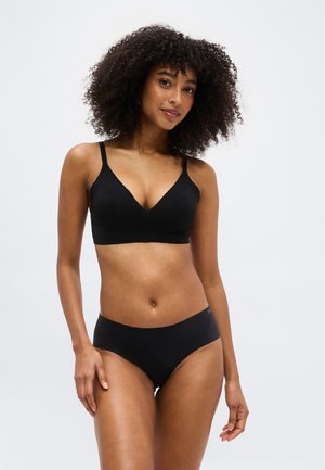 Frau mit lockigem Haar, die ein schwarzes Bralette und schwarze Unterwäsche trägt, steht vor einem schlichten hellgrauen Hintergrund.