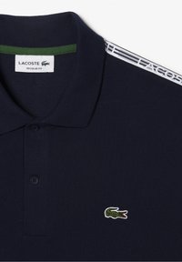 Marinblå Lacoste-pikétröja med klassisk krage, tre-knapps knäppning och en grön krokodil-logotyp broderad på bröstet.