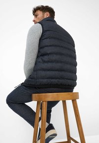 Marineblauer Steppweste mit hohem Kragen, getragen über einem grauen Pullover. Verfügt über gesteppte Paneele und einen kontrastierenden schwarzen Saum. Sitzt auf einem Holzschemel.