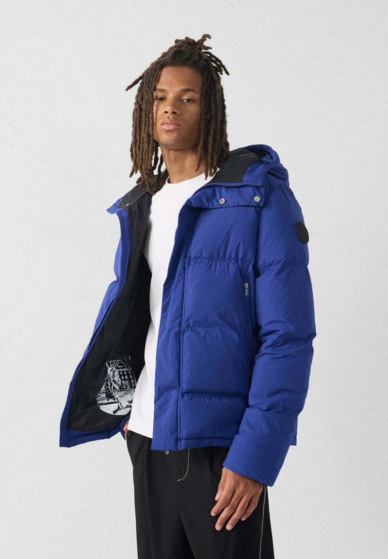 Blauwe geïsoleerde pufferjas met een rits- en knoopsluiting, voorzien van een capuchon en zwarte accenten, gedeeltelijk open geritst om het interieurpatroon te tonen.