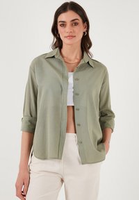 Světlonzelená košile na knoflíky s límečkem, vyhrnutými rukávy a uvolněným střihem, nošená přes bílý crop top a stylizovaná s neutrálními kalhotami.