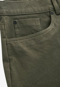 Olijfgroene broek met een gestructureerde stof, een voorzak met een zichtbare zwarte knoopaccent, en een gebogen tailleband detail.