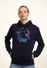 Giovane che indossa un hoodie blu navy con un mob tridimensionale pixelato a tre teste e il testo "TEMI IL WITHER!" sulla parte anteriore, che guarda leggermente in su e a destra.