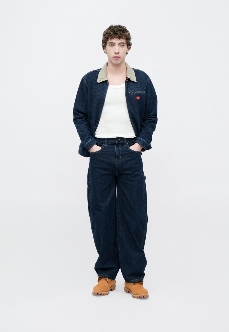 Veste en jean foncé avec col beige, associée à un pantalon ample en jean foncé. Débardeur blanc en dessous. Sabots marron complètent la tenue.