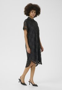 Black lace dress with short sleeves, button-down front, intricate floral pattern, and a curved hemline, paired with black heeled shoes.
