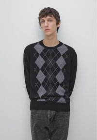Young man standing against plain wall wearing black sweater with gray argyle pattern and dark gray jeans, hands behind back.
