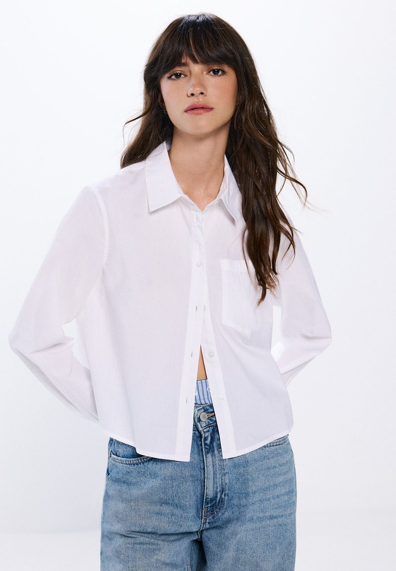 Camicia bianca a maniche lunghe con colletto e taschino sul lato sinistro del petto. Tessuto leggero, design cropped, abbinata a jeans in denim blu.