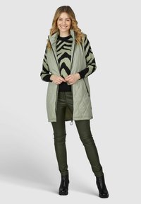 Helles grünes gesteppte Weste über einem schwarz-grünen zebraartigen Pullover, kombiniert mit olivgrünen Hosen und schwarzen Knöchelstiefeln.