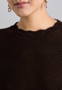 Brauner Strickpullover mit einem strukturierten Muster und einem gewellten Ausschnitt. Das Material wirkt leicht und atmungsaktiv. Ein silberner Ohrring ist sichtbar.