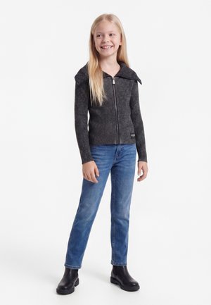 Ung pige med langt blondt hår iført en mørk ribstrikket lynlås-sweater, blå jeans og sorte ankelstøvler, smilende og stående.