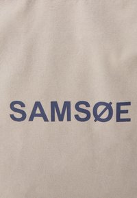 Borsa in tela beige con testo blu scuro che recita "SAMSØE." Presenta una superficie liscia e una forma rettangolare semplice.