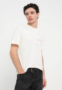 Βαμβακερό T-shirt εκρού με στρογγυλή λαιμόκοψη, κοντά μανίκια και μικρό κόκκινο κεντητό λογότυπο στο στήθος. Μαύρο τζιν που φοριέται με ζώνη.