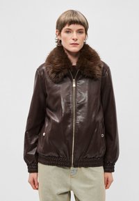 Brun læderjakke med pelskrave, elastiske manchetter og kant, samt en frontlynlås. Glat tekstur med sidelommer. Enkel design.